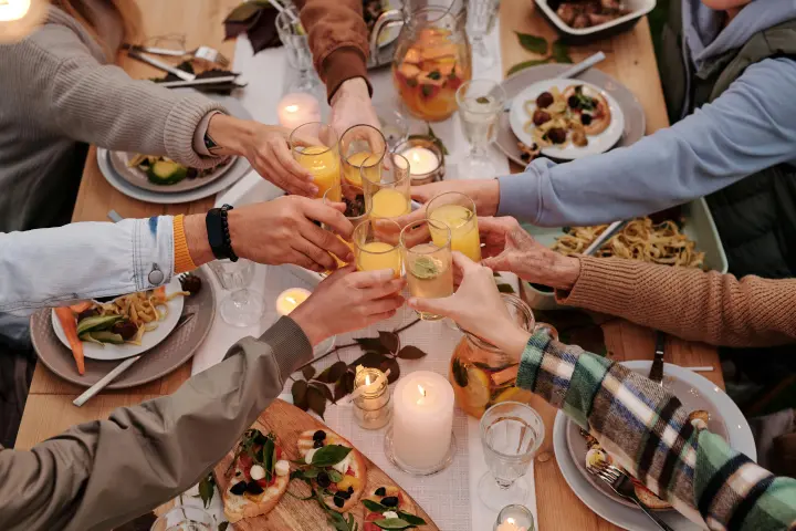 The hands of seven people reaching across a dinner table to clink glasses together for a toast. 