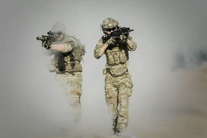 Two solders in full combat gear and guns at the ready are striding out of a cloud of smoke.