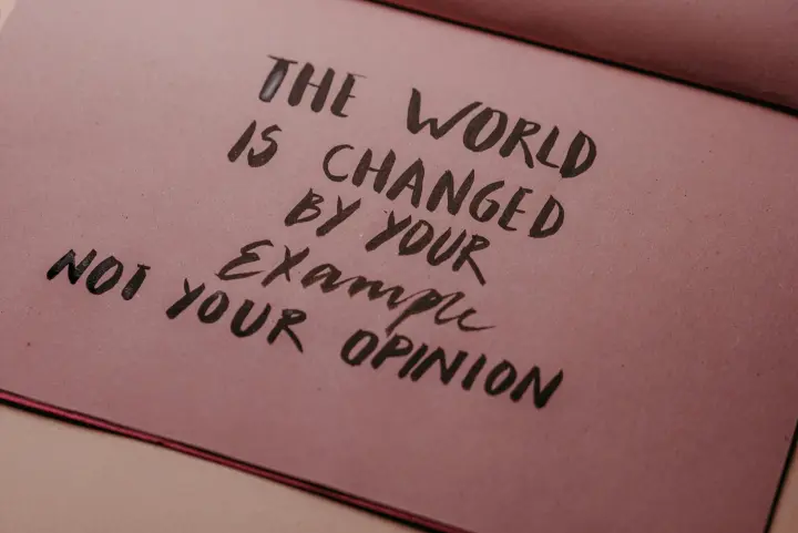 A brown card with black lettering, "The world is changed by your example not your opinion."