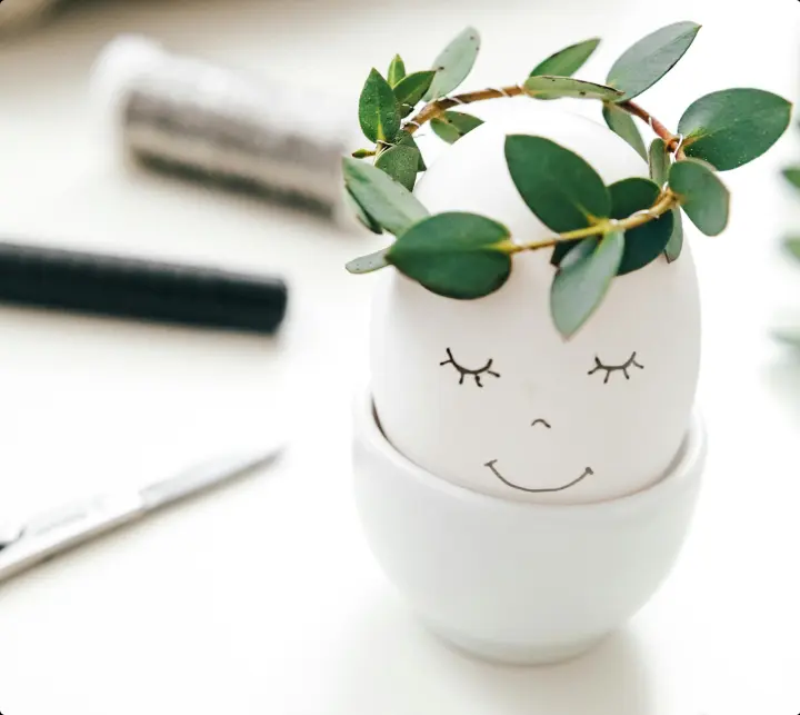 An egg with a smiling face and a sprig rosemary for a crown is sitting in an egg holder on a table.