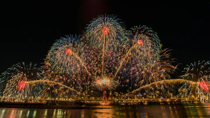 Time lapse capture of fireworks reflecting off the water of a shoreline.