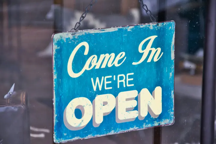 Blue sign with white letters "Come In We're OPEN" hanging in a store window.